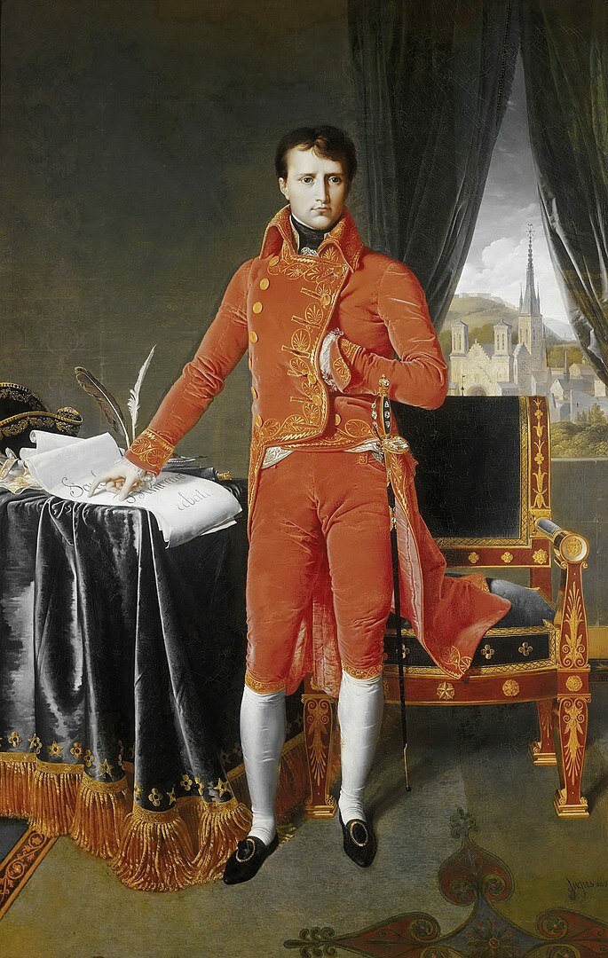 Kliknij, aby powiększyć Na obrazie portret Napoleona Bonaparte. Jest on ubrany w purpurowy uniform. Strój składa się ze spodni do kolan, białych podkolanówek i czarnych, wsuwanych butów ozdobionych klamrami. Żakiet mężczyzny ma duży podniesiony kołnierz, wyszywany jest złotą nitką we wzory z motywem liści. Żakiet z przodu jest krótki, do pasa. Ma dwa rzędy złotych guzików. Z tyłu - długi z rozporkiem, kończy się na wysokości kolan. Mankiety również wyszywane są złotą nitką. Cesarz lewą rękę ma schowaną na wysokości piersi za żakiet. Przy pasie z boku widać znajdującą się w pochwie szpadę, jej rękojeść wysadzana jest klejnotami. Prawa ręka Napoleona wskazuje na zapisany dokument, który leży na stole przykrytym grubym, aksamitnym, szarym materiałem zakończonym lamówką ze złotymi wzorami i frędzlami. Obok dokumentu kałamarz z zamoczonymi w nim dwoma piórami. Nieco dalej na stole widać fragment nakrycia głowy. Czapki dwurożnej czarnej, wykończonej złotymi haftami. Przed nakryciem głowy leżą białe haftowane rękawiczki. Z prawej strony obok cesarza stoi fotel, bogato zdobiony. Siedzisko i oparcie są z czarnej tkaniny. Elementy drewniane takie jak: nogi, podłokietniki są czerwone. Zdobienia fotela są złote. w pomieszczeniu na podłodze leży dywan z dużą rozetą pośrodku i złotym wzorem na brzegach. W oknie częściowo przesłoniętym ciemnymi kotarami widać panoramę Liège z katedrą św. Lamberta a dalej wzgórzami i nad nimi skłębionymi szarymi chmurami.