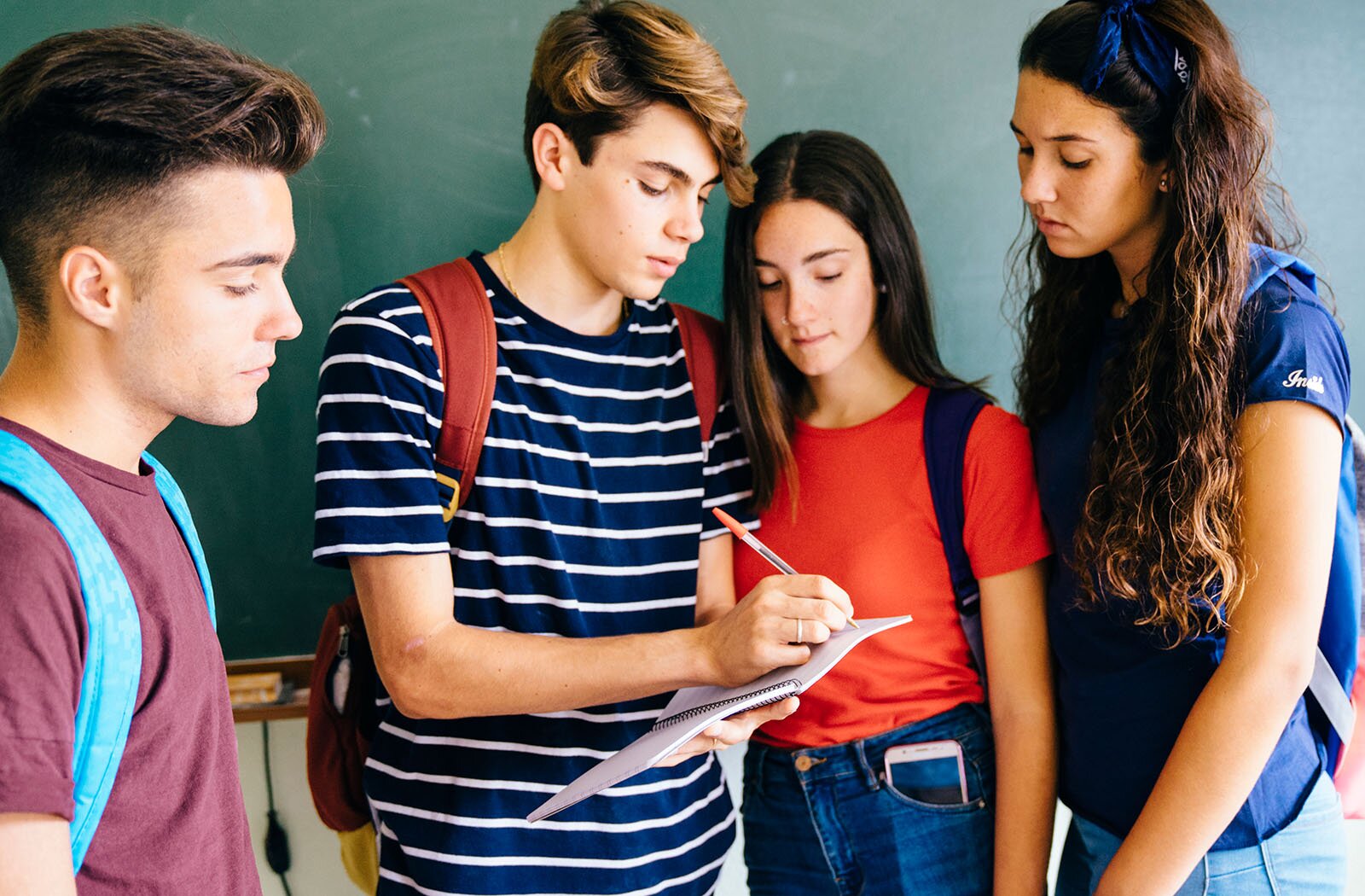 Zdjęcie przedstawia cztery młode osoby stojące obok siebie.  Od lewej widoczny jest młody chłopak, ustawiony bokiem, z krótkimi, ciemnymi włosami. Ubrany jest w bordową bluzkę z krótkim rękawem, a na plecach ma niebieski plecak. Obok niego stoi chłopak zwrócony frontalnie, z krótkimi, jasnymi włosami, głową skierowaną w prawo i lekko pochyloną. Ubrany jest w bluzkę z krótkim rękawem w biało-granatowe paski, a na plecach ma czerwony plecak. W lewej ręce trzyma otwarty zeszyt, a w prawej długopis, którym coś zapisuje.  Po jego prawej stronie znajduje się dziewczyna z długimi, ciemnymi włosami, pochylona i spoglądająca na zeszyt. Ubrana jest w czerwoną bluzkę z krótkim rękawem, a na plecach ma plecak. Ostatnia osoba to dziewczyna z długimi, ciemnymi włosami, stojąca lekko bokiem, zwrócona w lewo i również patrząca na zeszyt. Ubrana jest w granatową bluzkę z krótkim rękawem, a na plecach nosi niebieski plecak.  W tle widoczna jest zielona tablica szkolna.
