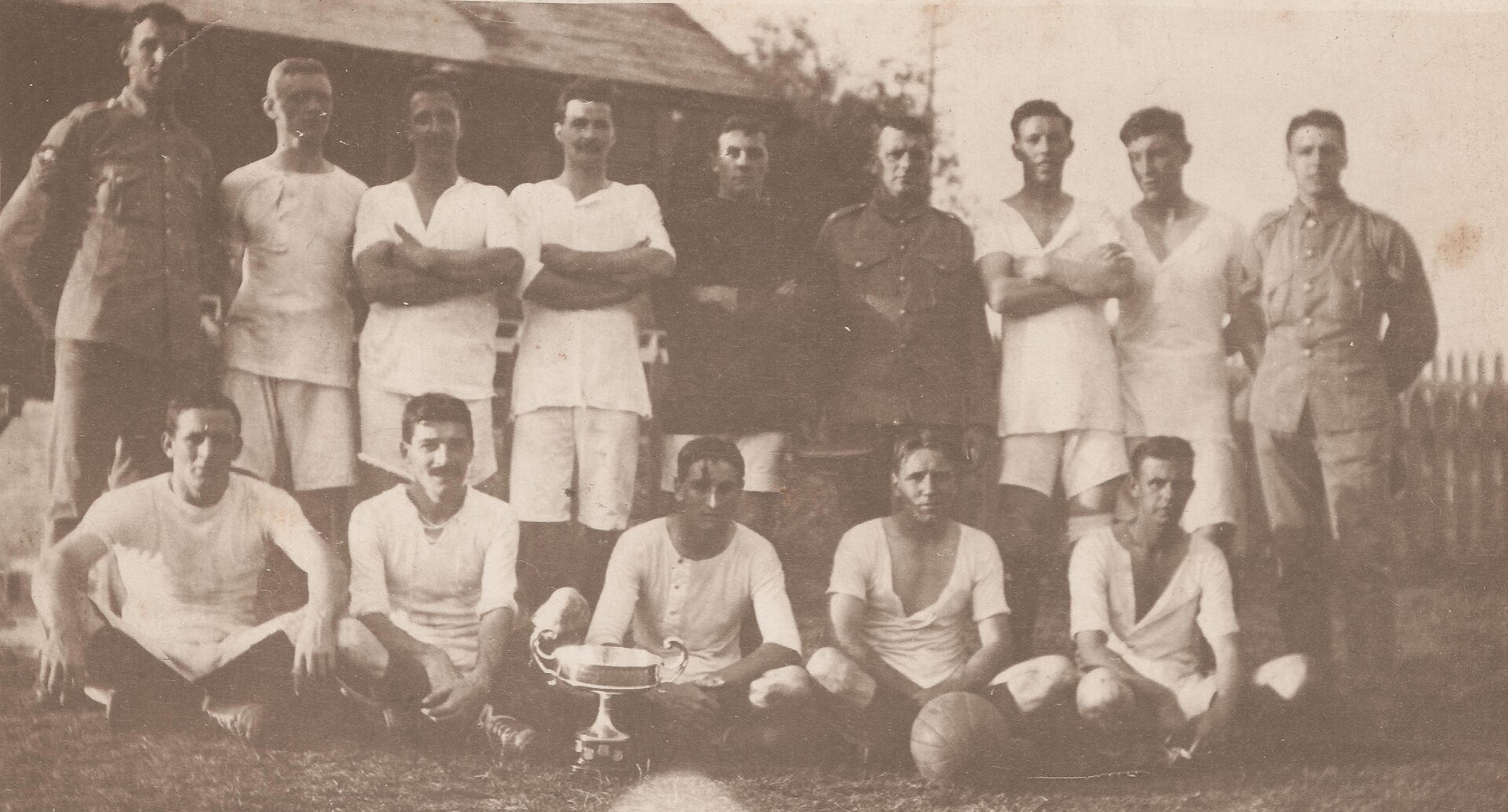 Zdjęcie przedstawia drużynę, która pozuje do zdjęcia. Składa się z dwóch rzędów młodych mężczyzn. Pierwszy rząd siedzi na trawie, a drugi stoi. Większość jest ubrana w jasne stroje sportowe, a kilku w mundury. Przed drużyną stoi puchar.