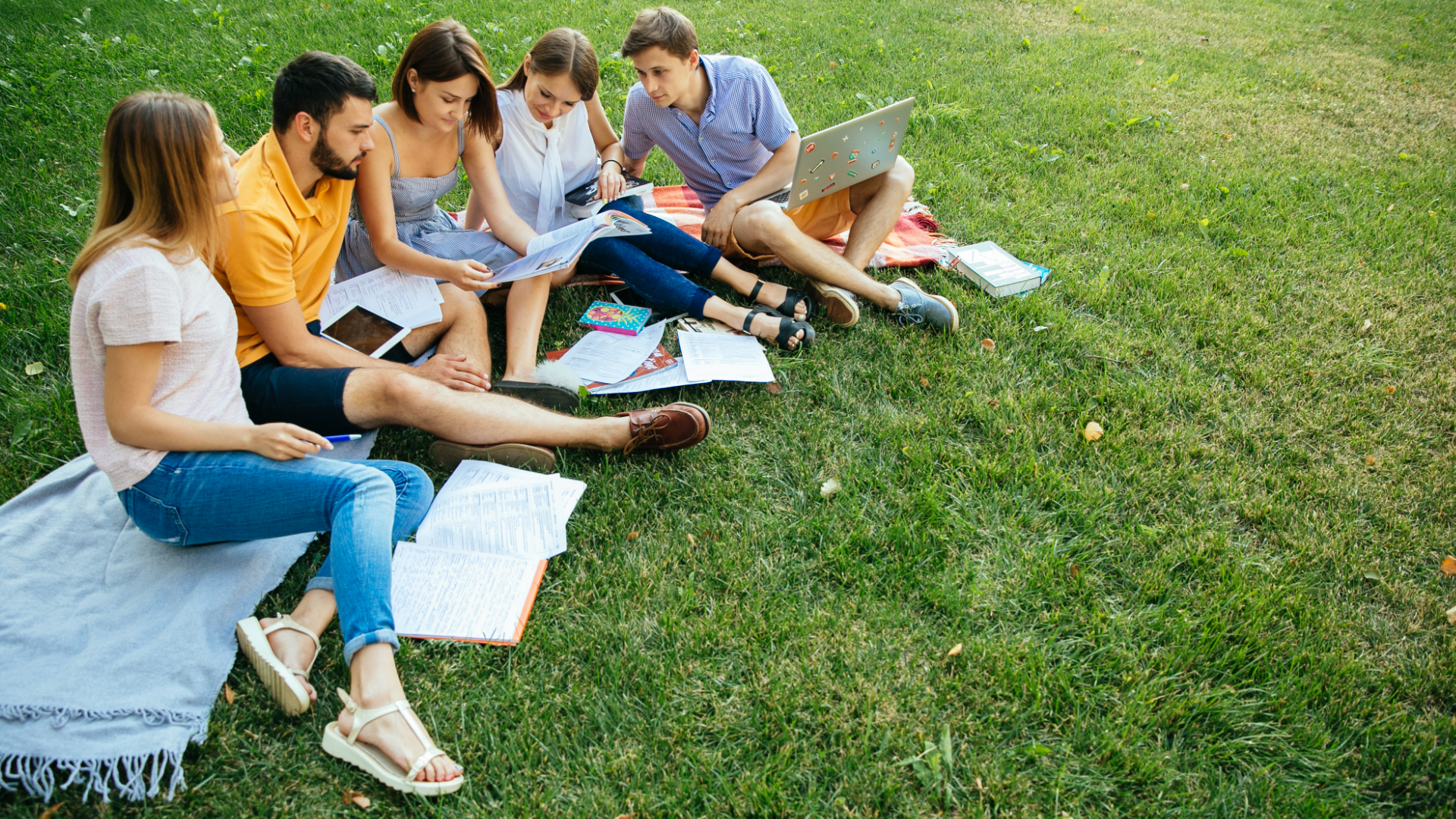 Zdjęcie przedstawia pięcioro nastolatków, trzy dziewczyny i dwóch chłopaków. Młodzież siedzi na kocu na trawie. Niektórzy mają przed sobą otwarte zeszyty, a jeden z chłopaków trzyma na kolanach laptopa. Młodzież razem się uczy.