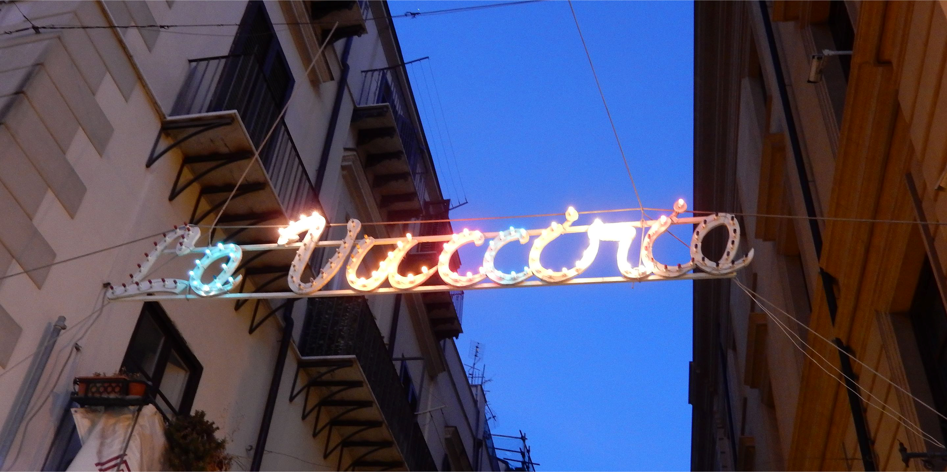 Zdjęcie przedstawia kolorowy neon z napisem La Vucciria na tle ciemnego nieba. Neon rozpięty jest pomiędzy dwiema kamienicami.