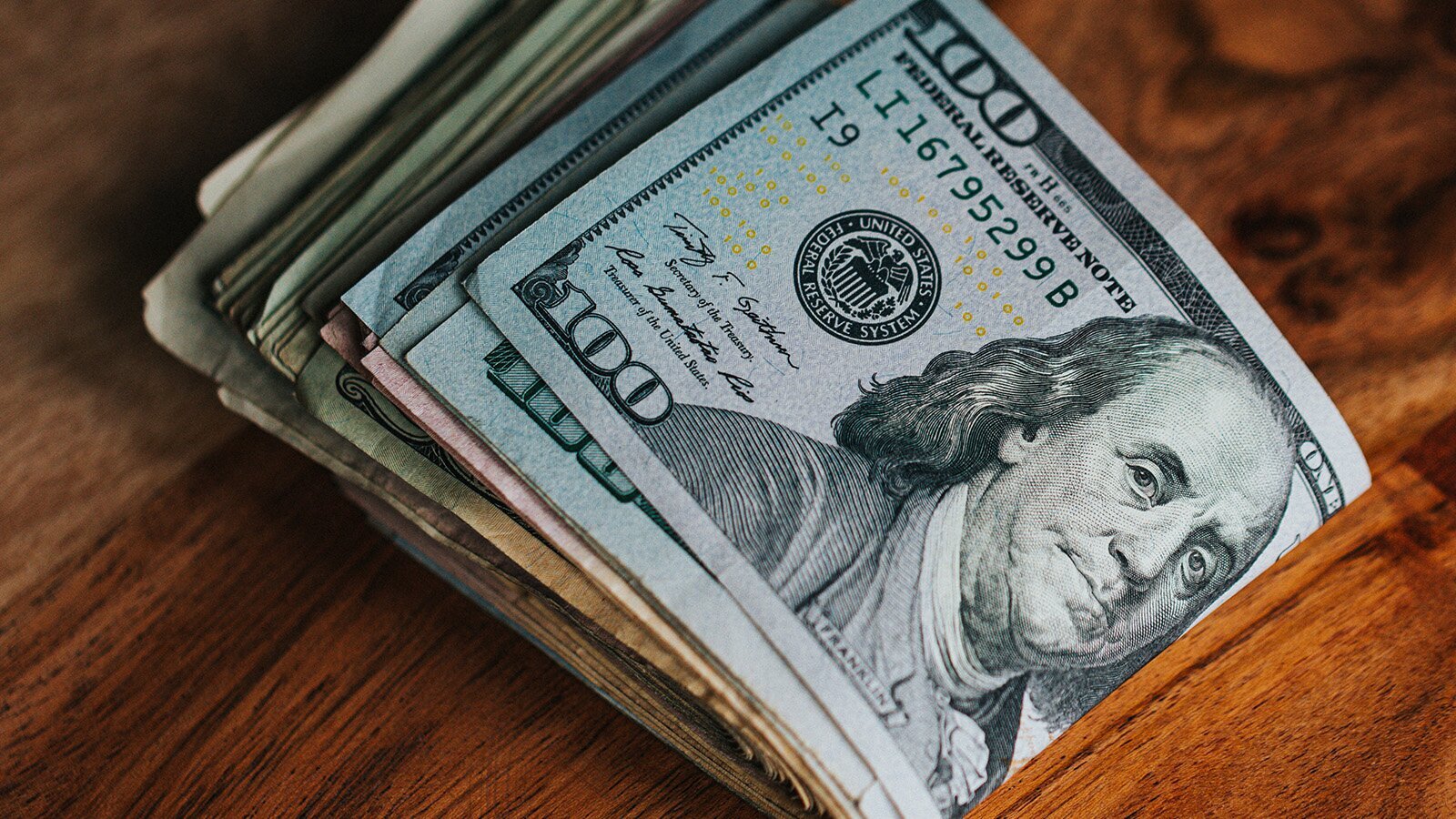 Zdjęcie przedstawia złożony na pół plik dolarów amerykańskich o różnych nominałach. Na pierwszym planie widoczny jest banknot o nominale 100, na którym znajduje się portret Benjamina Franklina z podpisem w języku angielskim: Franklin. Na lewo od niego widoczny jest okrągły stempel z dookolnym napisem w języku angielskim: UNITED STATES FEDERAL RESERVE SYSTEM. Powyżej widoczny jest napis w języku angielskim: FEDERAL RESERVE NOTE oraz numer seryjny, a poniżej widoczne są dwa podpisy. Pod pierwszym widać napis w języku angielskim:  Secretary of the Treasury,  a pod drugim: Treasurer of the United Sta Treasurer of the United States. 