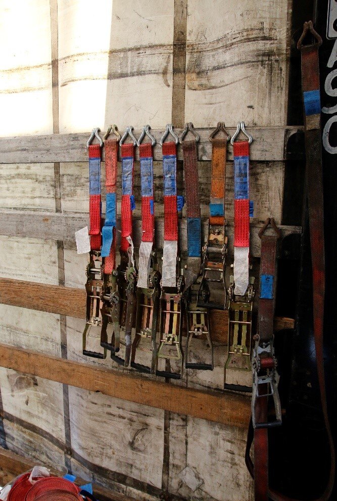 Ilustracja przedstawia dziewięć pasów mocujących. Osiem jest w kolorze czerwonym jeden w pomarańczowym, pasy są w różnym stanie, na większości z nich widać ślady eksploatacji. Każdy pas posiada klamrę. Składają się one z jednej części pasa, którego koniec zakończony jest specjalna klamrą zaciskową.