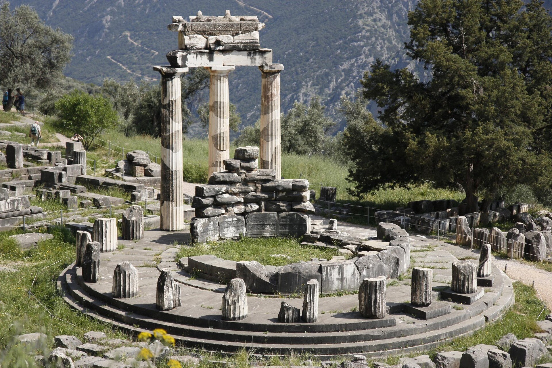 Zdjęcie przedstawia wzgórze, na którym znajdują się wpisane w okrąg ruiny starożytnej świątyni. Widoczne są pozostałości kolumn otaczające świątynię oraz ułożone z bloków kamiennych resztki jej ścian.