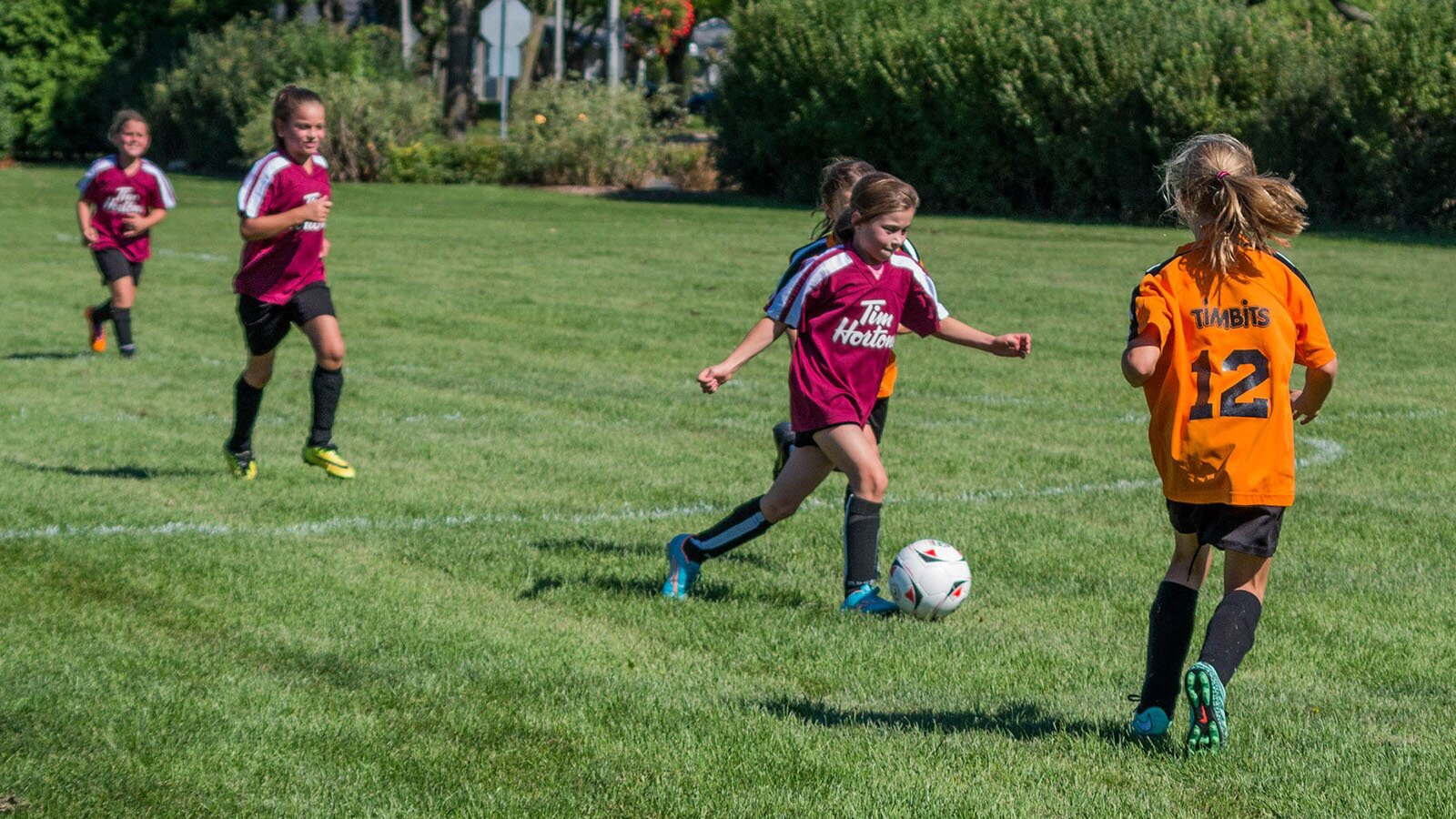 Zdjęcie przedstawia dziewczynki grające w piłkę nożną na murawie piłkarskiej. Wszystkie ubrane są w stroje piłkarskie. Na przodzie widoczna jest dziewczynka grająca na pozycji pomocnika, biegnąca w kierunku piłki. Naprzeciw niej biegnie dziewczynka z drużyny przeciwnej. 