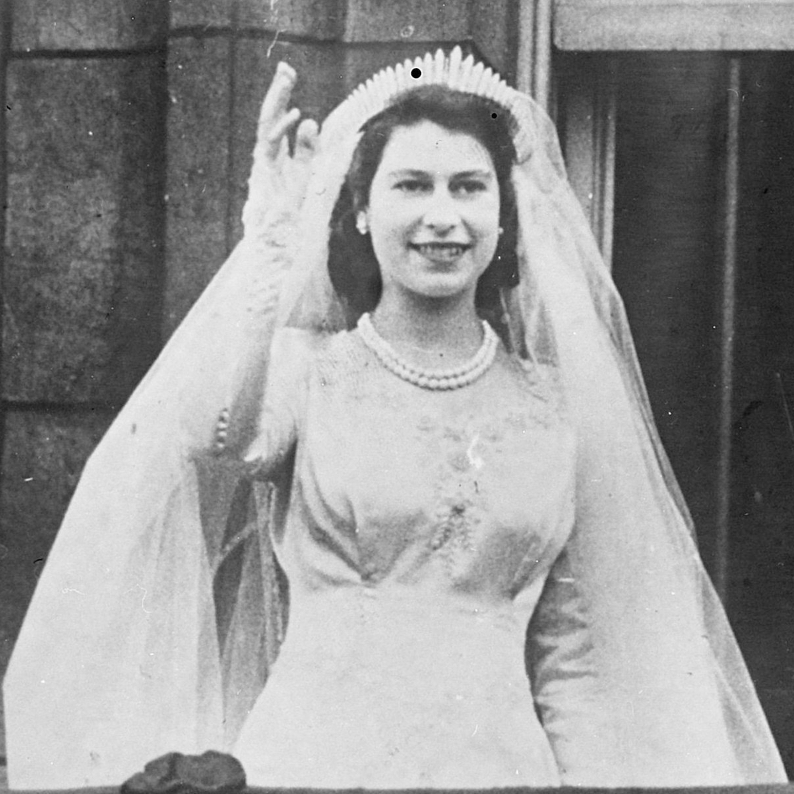 Czarno‑białe zdjęcie przedstawia młodą królową Elżbietę II w trakcie ślubu. Widoczna jest młoda dziewczyna, w ujęciu od pasa w górę, ubrana w białą suknię i welon, z naszyjnikiem pereł na szyi. Dziewczyna ma podniesioną do góry prawą rękę, a palce dłoni są lekko rozchylone i układają się w geście pozdrowienia. Dziewczyna uśmiecha się.