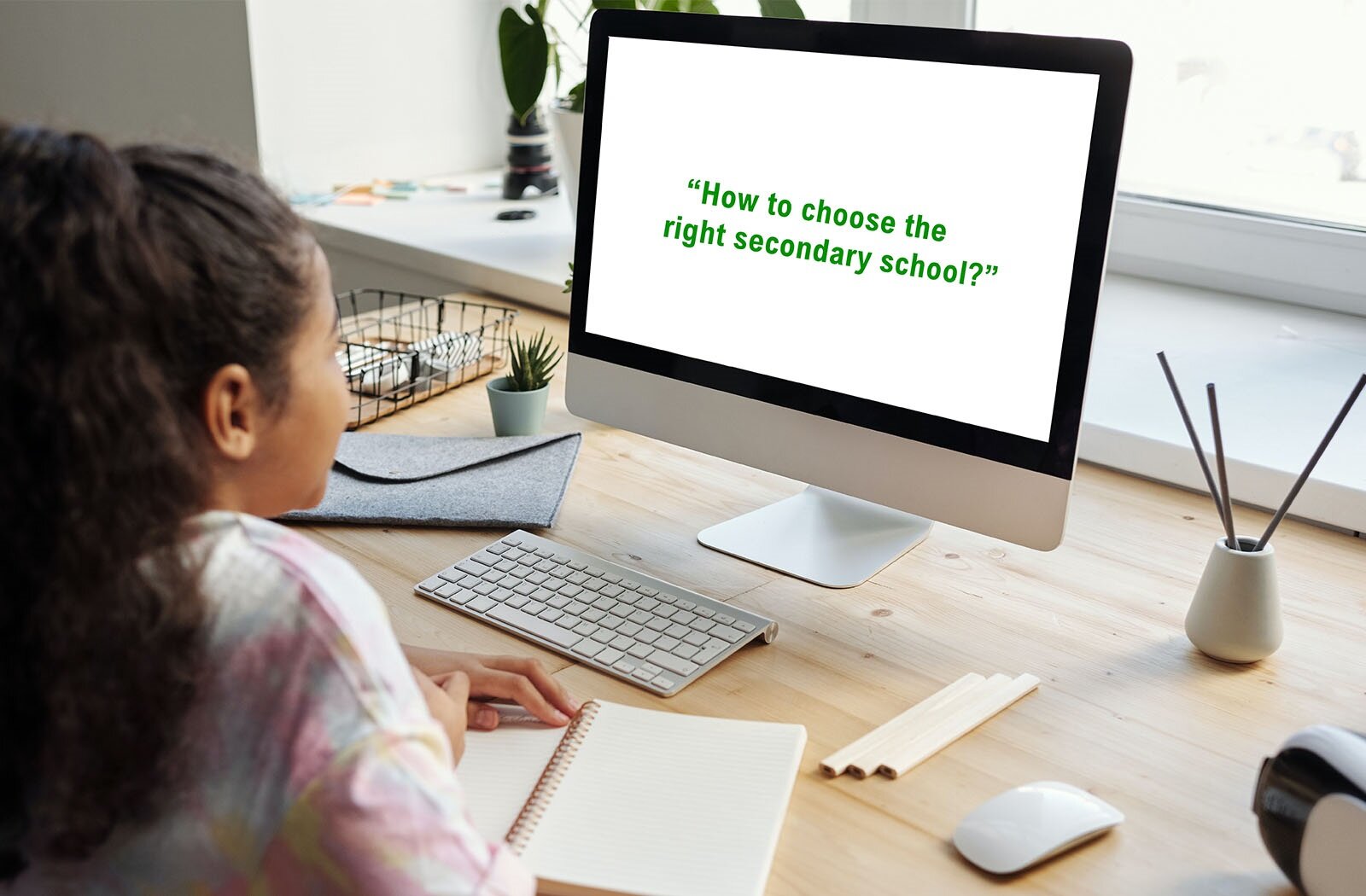 Zdjęcie przedstawia nastolatkę w ujęciu z tyłu, siedzącą przy biurku. Jest ubrana w kolorową bluzkę i ma długie, ciemne włosy związane w kucyk. Spogląda na monitor komputera. Jej dłonie spoczywają na otwartym notatniku.  Na biurku stoi monitor, na którego ekranie widoczny jest napis w języku angielskim: "HOW TO CHOOSE THE RIGHT SECONDARY SCHOOL?". Obok znajdują się klawiatura, myszka, ołówki oraz pojemnik na dokumenty.
