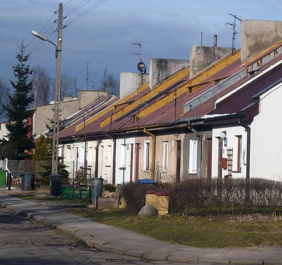 Zaniedbane blokowisko z kolorowymi dachami (żółte, czerwone, niebieskie) na peryferiach polskiego miasta, prawdopodobnie Łodzi; szare ściany z odpadającym tynkiem, śmietniki, słupy latarni i przewody elektryczne, otoczone chwastami i świerkami na tle pochmurnego nieba.