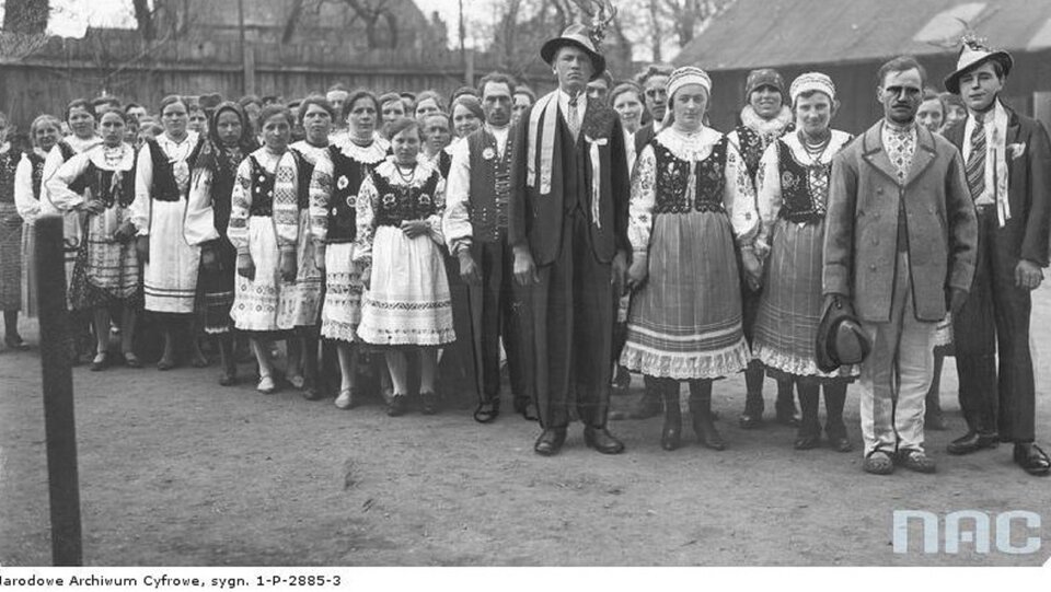 Ilustracja przedstawia liczną stojącą grupę Łemków ubranych w tradycyjne stroje ludowe. Kobiety ubrane w koszule z lnianego płótna, zwana „oplicza”. Przy szyi  przyszyte marszczone i nakrochmalone krezy zakończone ząbkami, ozdobione szlakiem białego haftu. Jedynym zapięciem jest tasiemka wiązana pod szyją. Do spódnicy noszono zapaskę, zdobioną kolorowym haftem. Mężczyźni w koszulach z płótna. Na przodzie proste rozcięcie, pod szyją wiązana tasiemka. Łemkowie nosili trzy rodzaje spodni: „gaczy”, „nohawky” i „chołoszni”. Uzupełnieniem męskiego stroju jest pas skórzany oraz kapelusz „uherski” - czarny, filcowy, otoczony kolorową wstążką.