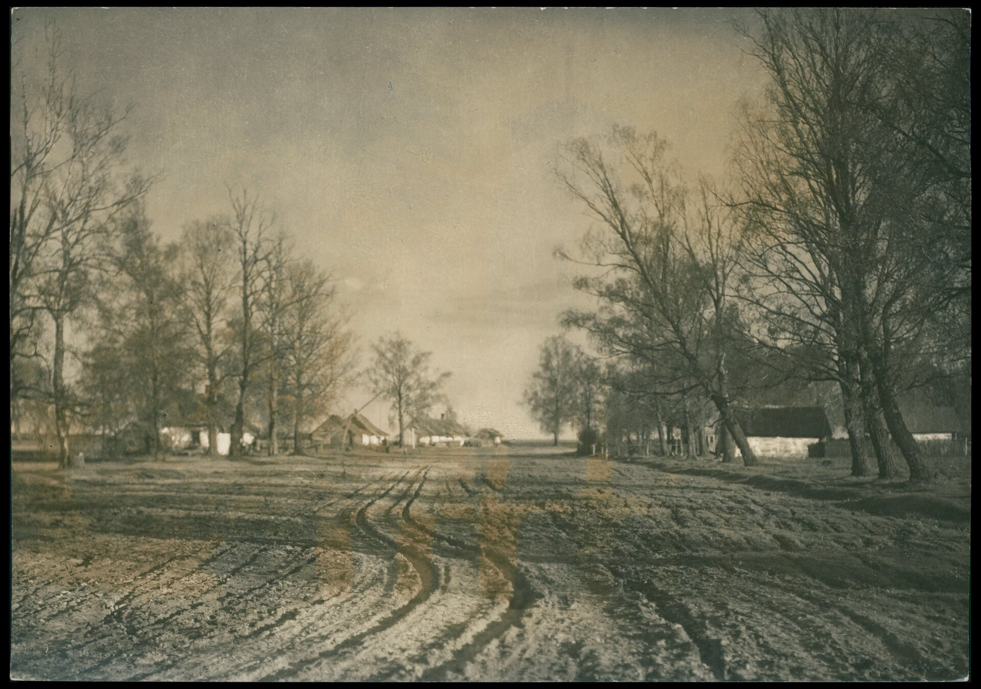 Stara, czarno‑biała fotografia przedstawiająca miejscowość ze starymi chatami. Zdjęcie wycentrowane jest na szeroką, piaszczystą drogę. Po obu stronach drogi drzewa i między nimi rozporoszone stare chaty.