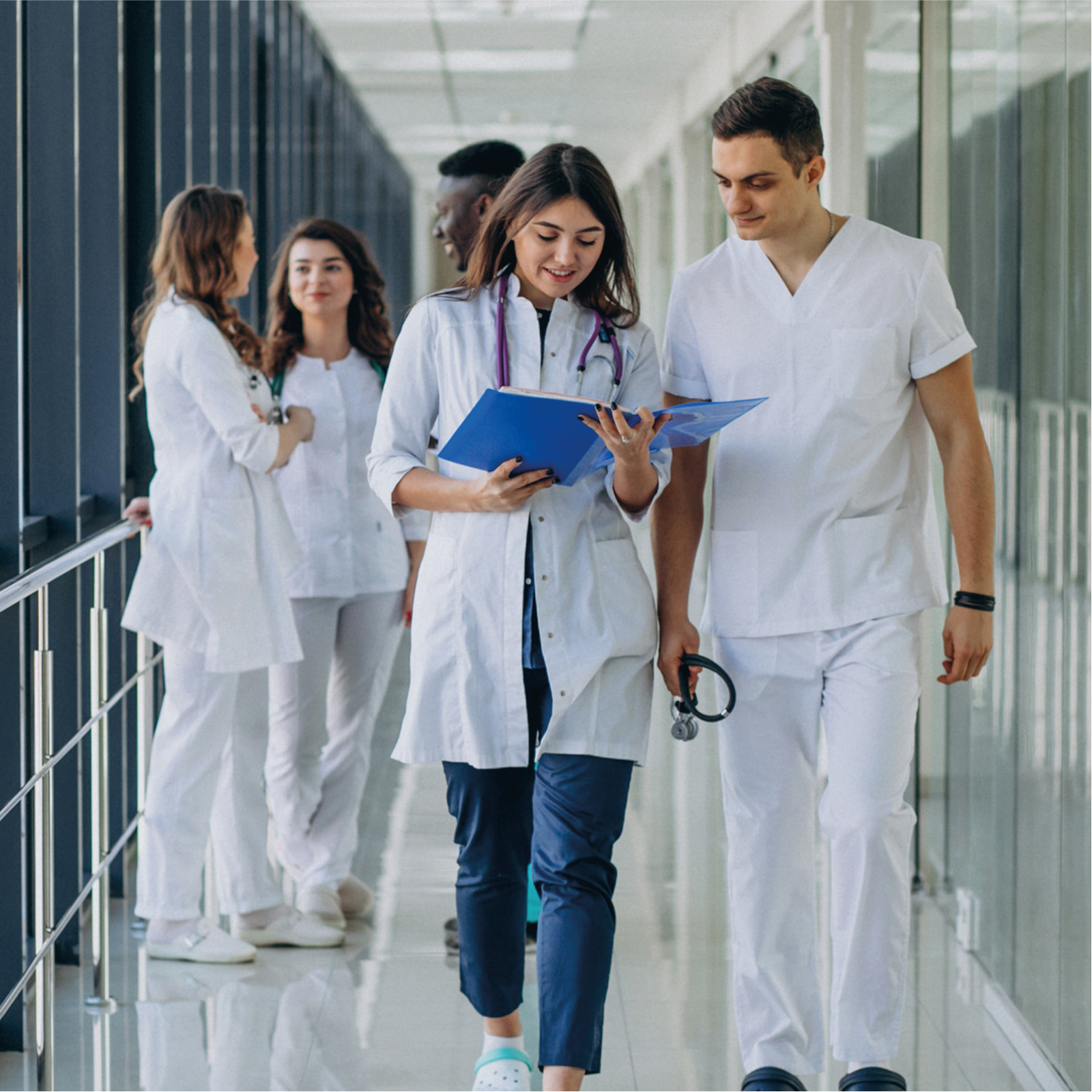 Zdjęcie przedstawia pięć osób w białych fartuchach i ze stetoskopami na szyjach. Troje z nich rozmawia na drugim planie, dwoje przechadza się przeglądając dokumentację medyczną.