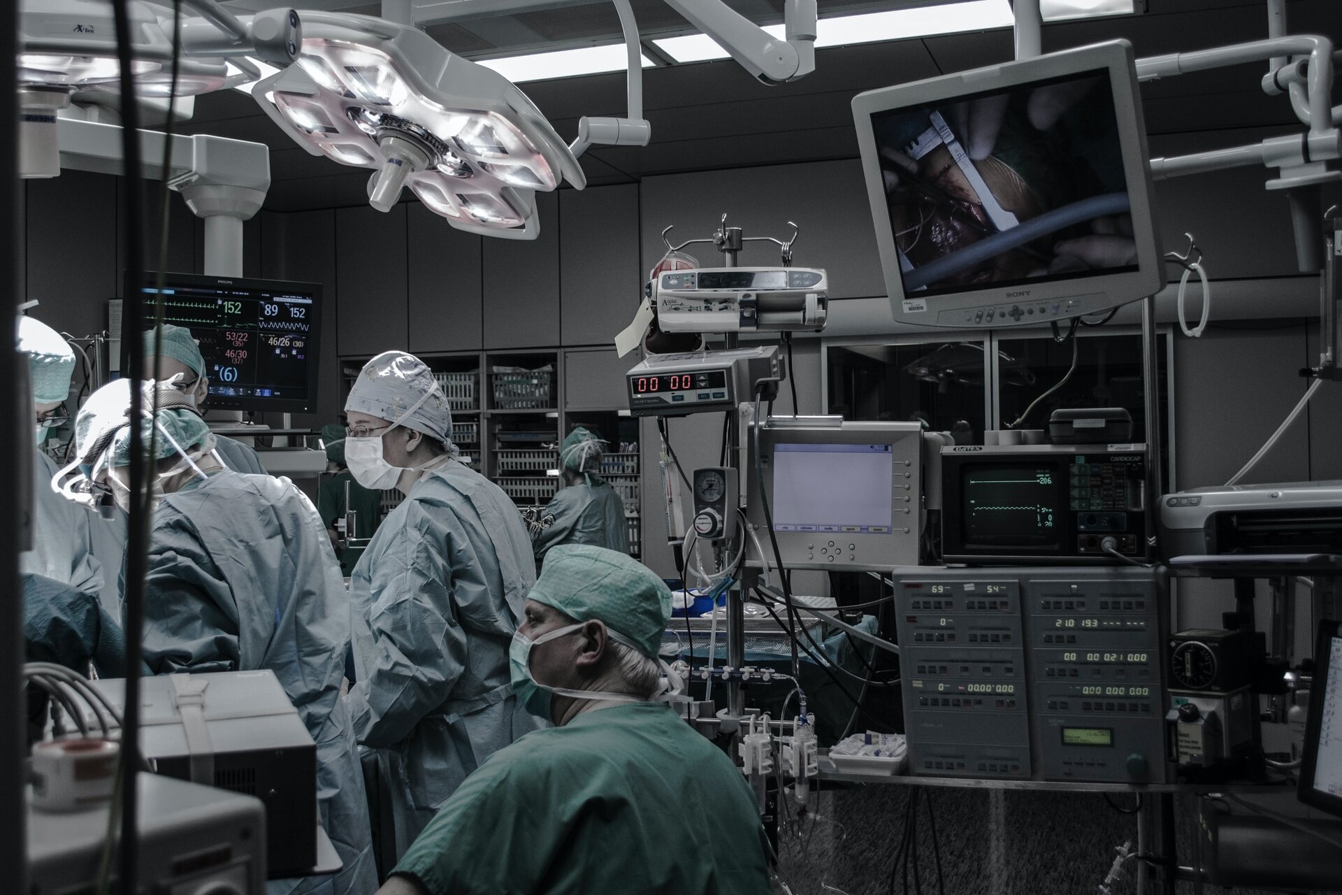 Fotografia przedstawiająca wnętrze sali operacyjnej. Lekarze ubrani w jednorazowe fartuchy, na głowach mają jednorazowe czepki, na twarzach maseczki chirurgiczne. W sali stoi sprzęt medyczny, u góry włączona lampa chirurgiczna. Wkoło pełno sprzętu medycznego, liczne monitory.