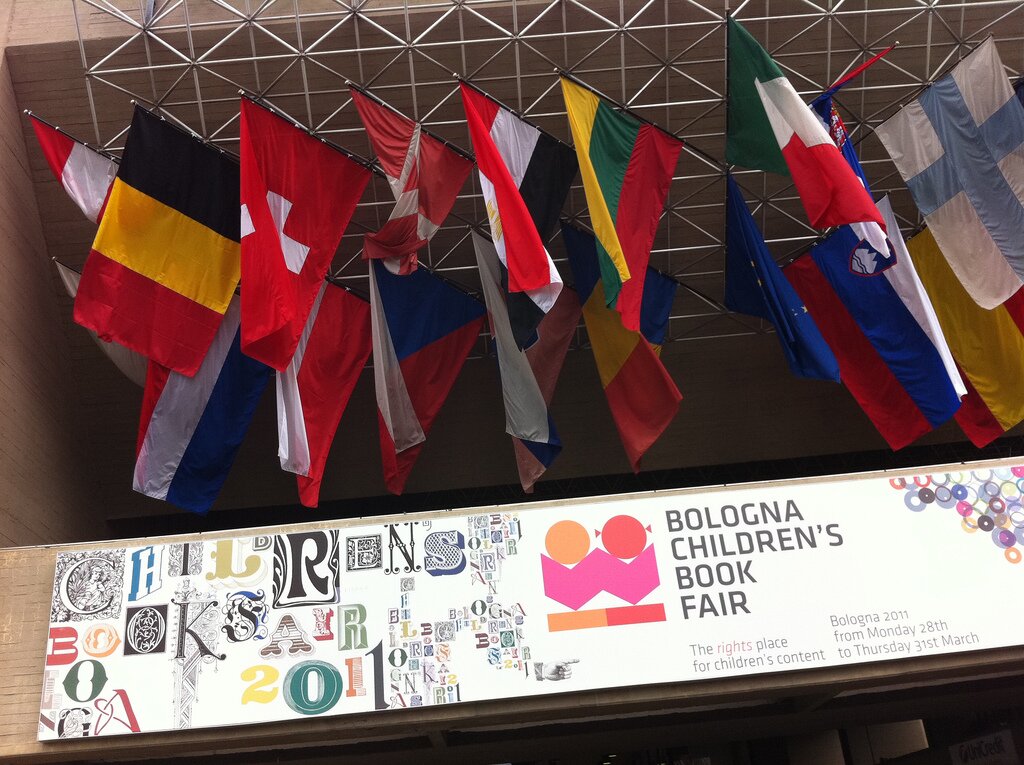 Ilustracja przedstawia flagi różnych krajów zawieszone na metalowym rusztowaniu na poddaszu. Pod flagami znajduje się informacja w języku angielskim.