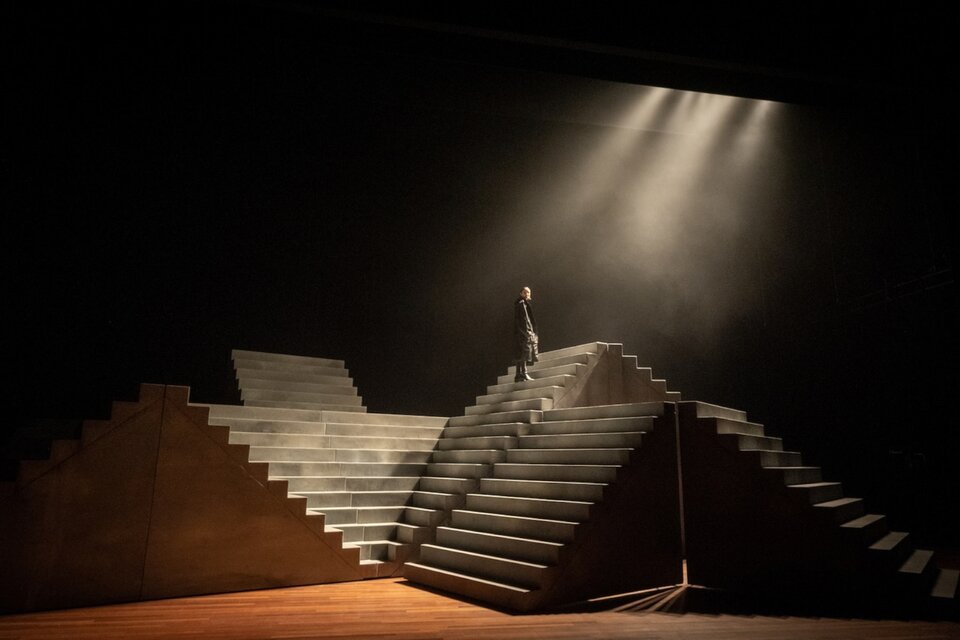 Fotografia przedstawia scenografię ustawioną na deskach teatru. Scenografia składa się z konstrukcji, na którą składają się schody ustawione obok siebie oraz jedne na drugich. Z prawej strony, na schodach stoi postać ubrana w ciemny płaszcz.