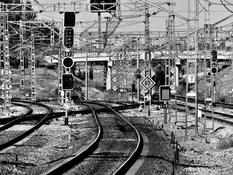 Zdjęcie przedstawia trzy tory kolejowe. Biegną one obok siebie. Między torami stoją słupy trakcji elektrycznej. Są także sygnalizatory świetlne i znaki kolejowe. Na drugim planie widoczny jest most stojący nad torami.