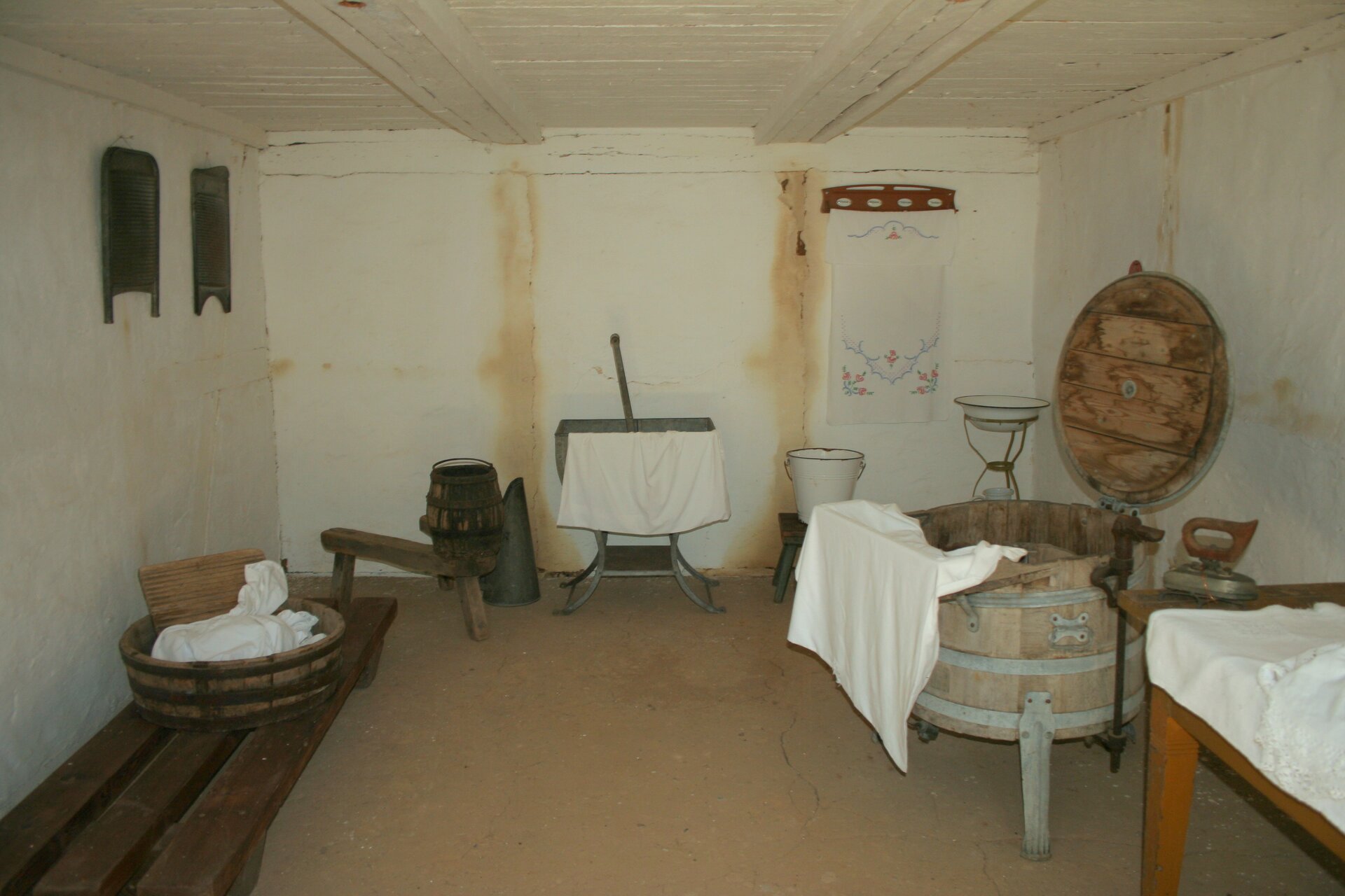 Zdjęcie przedstawia izbę gospodarczą w Muzeum Wsi Słowińskiej w Klukach. W pomieszczeniu ukazane są eksponaty, z których żyjąca wcześniej ludność korzystała, rzeczy takie jak sprzęt do prania ubrań z tarą w środku drewnianej miski, wiadra, balie do prania, żelazko i magiel. 