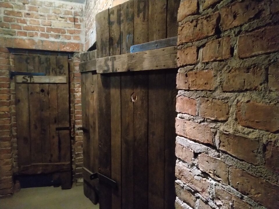 Ilustracja przedstawia ścianę murowaną z czerwonej cegły z trojgiem drewnianych drzwi. na pierwszych napisano czarną farba: Cela nr 1. 