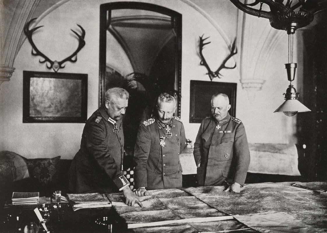Zdjęcie przedstawia trzech starszych mężczyzn w wojskowych mundurach, którzy stoją przy stole. Na ich mundurach wiszą medale i odznaczenia. Na stole rozłożone są mapy. Mężczyźni oglądają je, jeden wskazuje na jakiś fragment palcem. Za mężczyznami na ścianie wiszą obrazy oraz jelenie rogi.