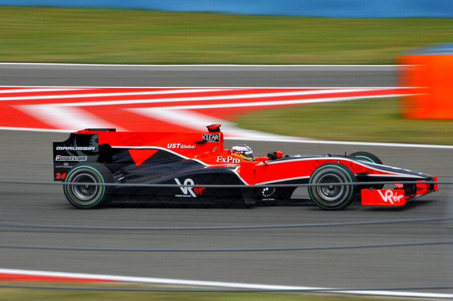 Zdjęcie przedstawia bolid Formuły jeden uchwycony z boku w ruchu z silnie rozmytym tłem. Zbliżenie na centralną część maszyny i kierowcę.