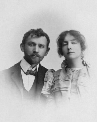 Czarno‑białe zdjęcie portretowe przedstawia kobietę i mężczyznę. Kobieta jest ubrana w sukienkę z długim rękawem. Ma upięte włosy. Mężczyzna jest ubrany w koszulę i garnitur. Pod szyją ma muszkę. Para stoi obok siebie. Oboje są poważni.