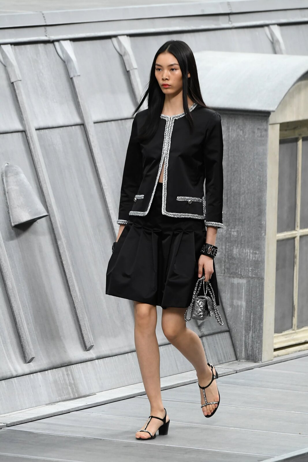 Fotografia przedstawia modelkę na wybiegu, mającą na sobie kreację Chanel. Jest to czarna marynarka ze srebrnymi detalami na brzegach, zapięta od góry tylko do połowy, czarna, luźna spódnica sięgająca przed kolano, srebrna, mała torebka oraz czarne sandały na niskim, grubym obcasie ze srebrnymi detalami. Na lewej dłoni kobiety widoczna jest gruba bransoleta, z materiału przypominającego czarną skórę, z elementami srebrnych liter. Usta kobiety pomalowane są na delikatny czerwony kolor, a jej czarne, proste włosy, opadają na jej dekolt i ramiona. 