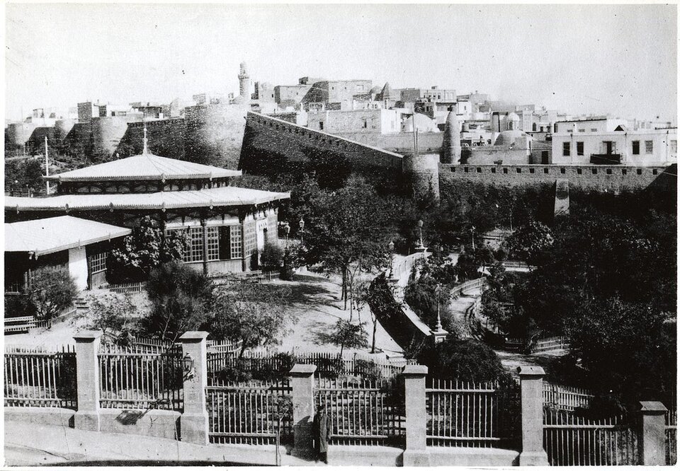 Czarno‑biała, archiwalna fotografia przedstawia widok na fragment miasta. W tle wznosi się wysoki mur obronny, za którym widoczna jest gęsta, historyczna zabudowa miejska z wieżą.