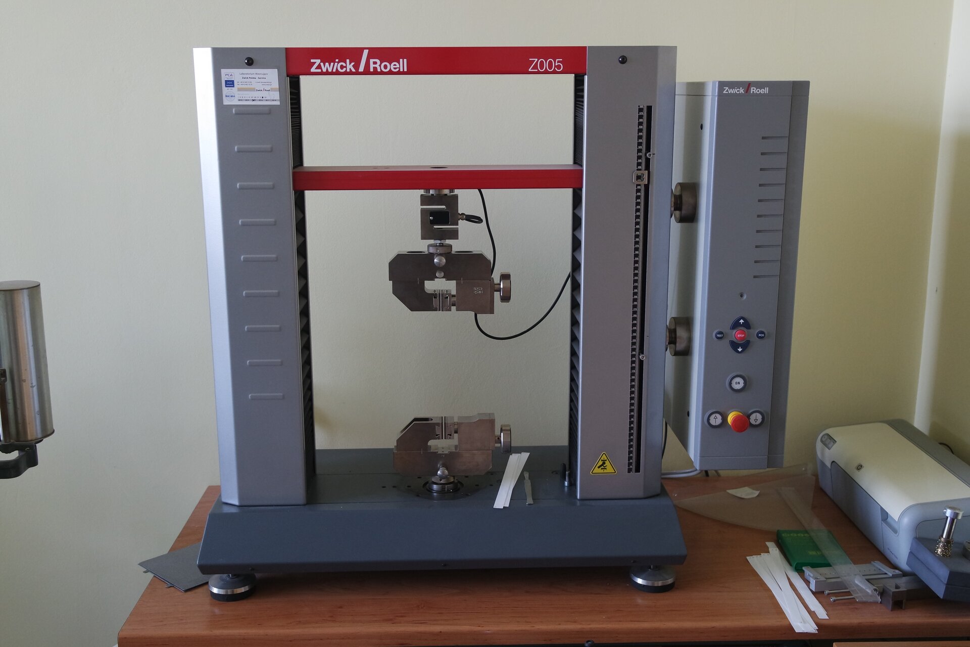 Grafika przedstawia zrywarkę pionową do oznaczania siły zrywania, samozerwalności, rozciągliwości. Urządzenie zostało umieszczone na blacie, składa się z dwóch szarych kolumn z czerwonymi łącznikami. Po prawej stronie widnieje panel sterujący.