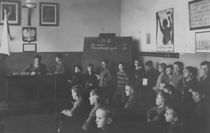 Fotografia przedstawia klasę w jednej ze szkół. Widać nauczyciela siedzącego przy biurku, wpisującego oceny. W  do biurka stoi kilkunastu chłopców reszta siedzi w ławkach. Nad biurkiem wisi godło i flaga Polski. Jeszcze wyżej Krzyż z Chrystusem. Po prawej stronie stoi tablica. Na ścianach widać obrazki.