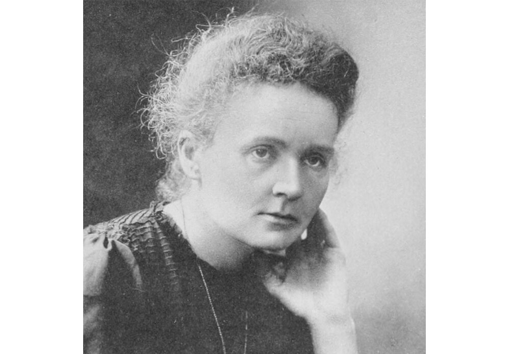Czarno-biała fotografia przedstawiająca Marię Curie-Skłodowską. Patrzy na coś poza kadrem. Ma na sobie ciemny strój, kręcone włosy są spięte. Jedną dłoń przykłada do policzka.