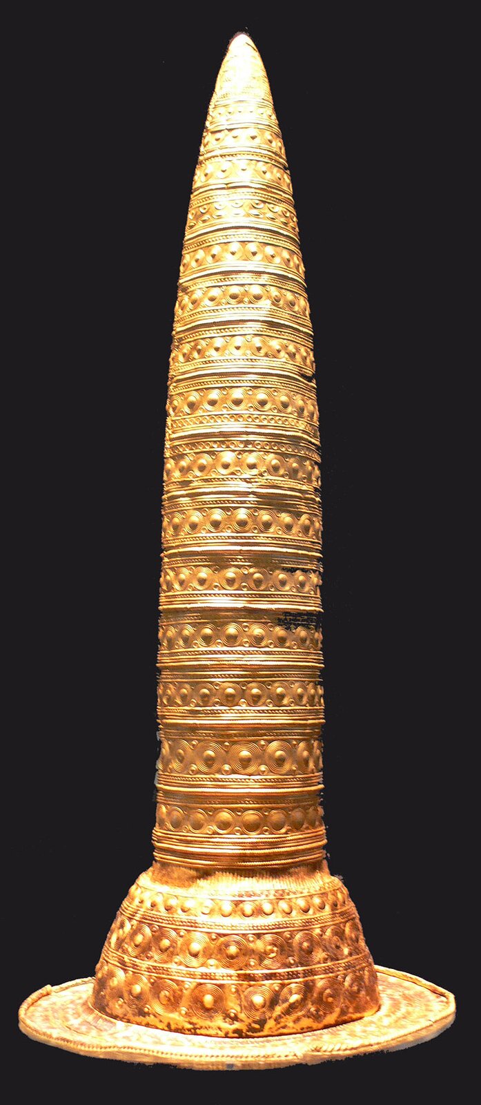 Zdjęcie przedstawia eksponat muzealny, którym jest złoty przedmiot w kształcie stożka, ale o niewielkiej różnicy średnicy na całej swojej długości. Przedmiot jest zdobiony geometrycznym ornamentem. 