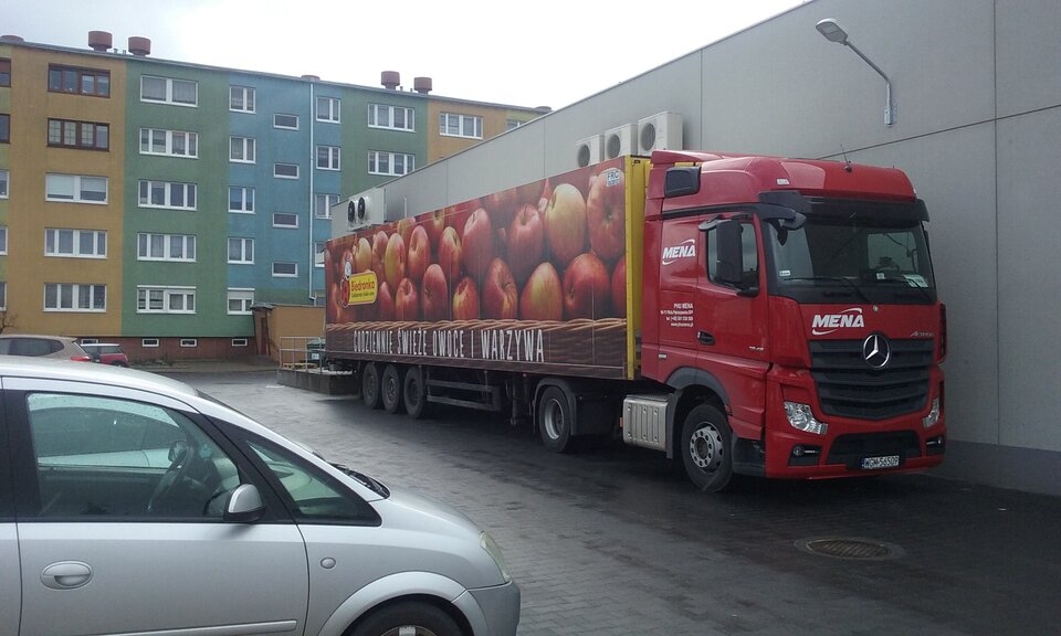 Zdjęcie przedstawia tira, który stoi przy ścianie dużego sklepu. Na plandece naczepy tira namalowane są jabłka. Za tirem i sklepem stoi blok. 