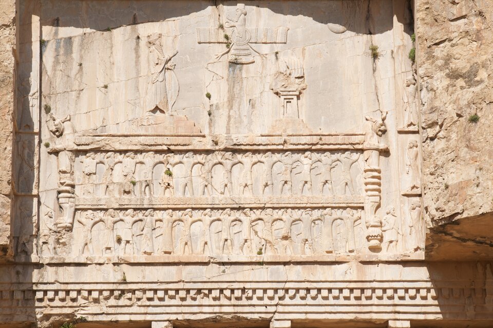 Ilustracja przedstawia fragment reliefu z grobowca. Na reliefie przedstawione zostały dwa rzędy postaci są to mężczyźni stojący w dwóch rzędach, trzymający się za ręce. Ponad nimi znajduje się fragment płaskorzeźby postaci.