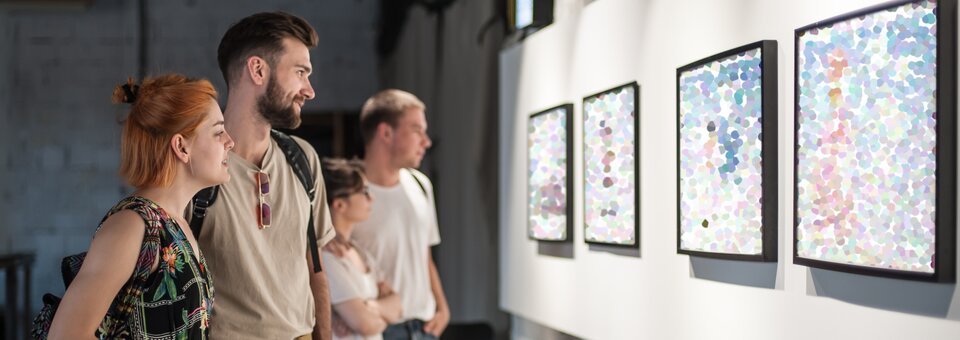 Zdjęcie przedstawia kilka osób oglądających wystawę prac. Najbliżej stoi młoda kobieta o rudych włosach i młody mężczyzna z zarostem. Cztery prace wiszą na białej ścianie, obok siebie. W czarnych ramkach znajdują się różnokolorowe kropki.