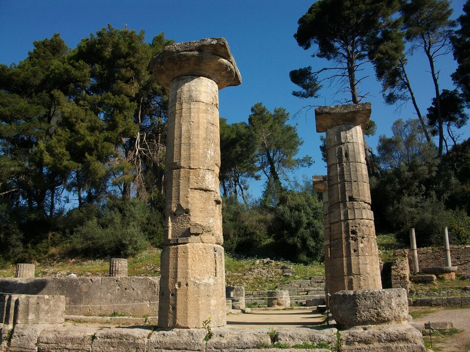 Ruiny Świątyni Hery w Olimpii, widoczne dwie kolumny, za nimi iglaste drzewa i niebieskie niebo.