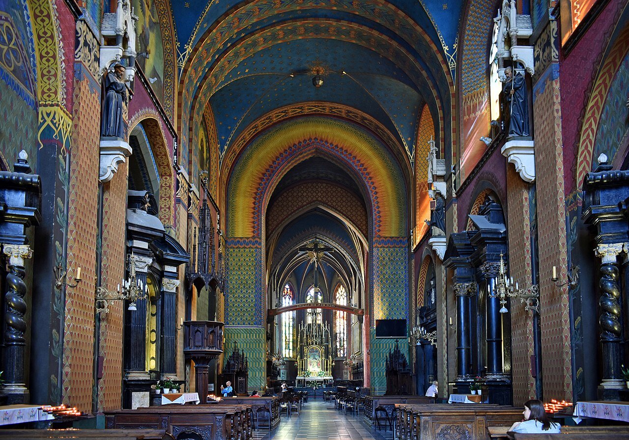 Ilustracja przedstawia bardzo długie wnętrze kościoła o łukowatym sklepieniu. Ostre łuki znajdujące się w elementach konstrukcyjnych oraz sklepienie krzyżowo-żebrowe, a także okna wypełnione witrażami.