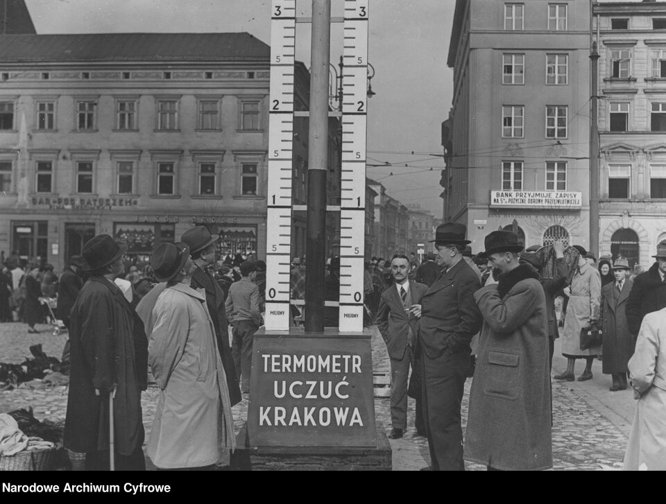 Zdjęcie przedstawia konstrukcję termometru z podpisem TERMOMETR UCZUĆ KRAKOWA. Instalacja stoi na miejskim placu. Wokół niej są zebrani ludzie.