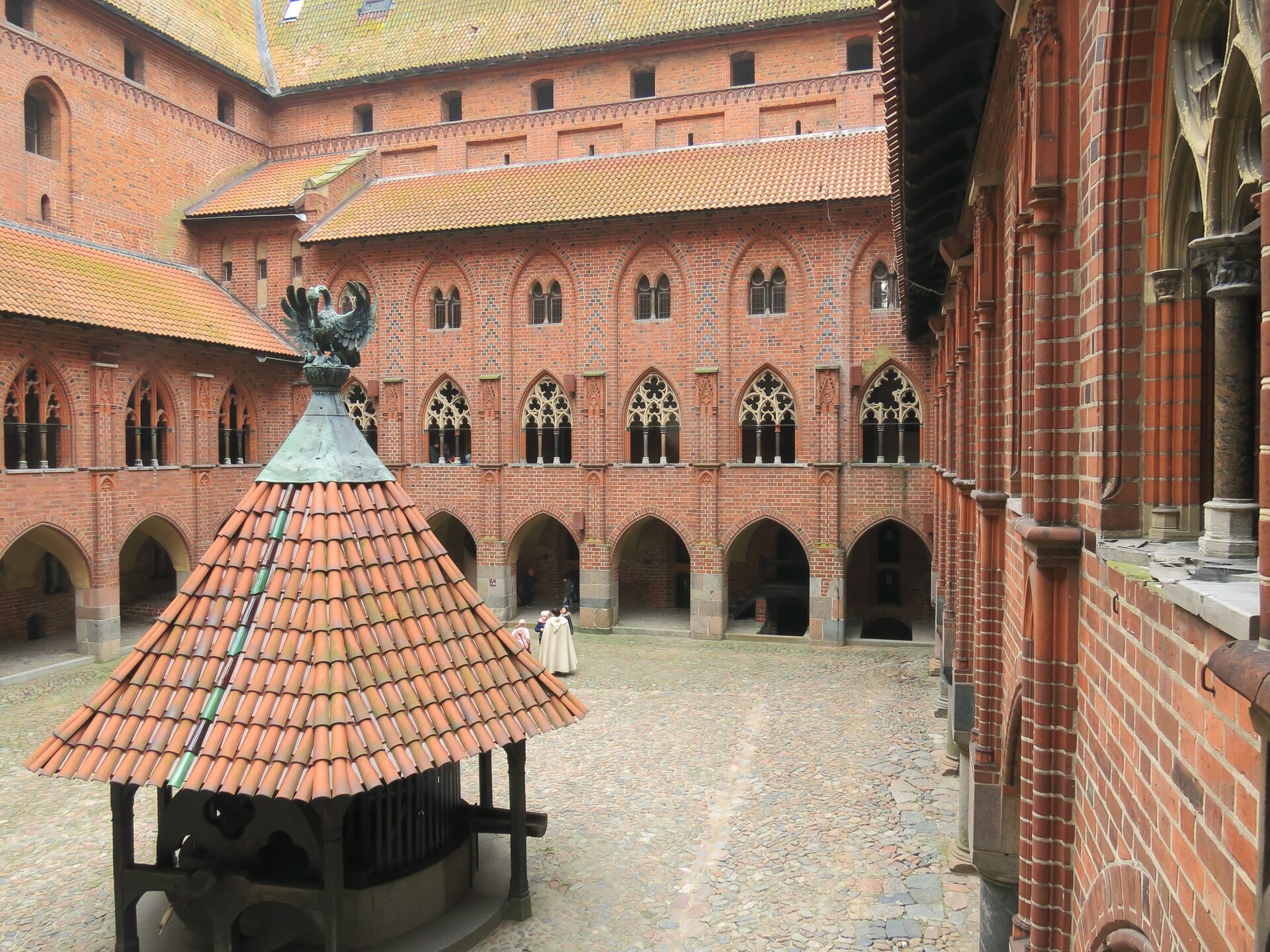 Zdjęcie przedstawia fragment dziedzińca zamku w Malborku. Po środku dziedzińca stoi zadaszona studnia.