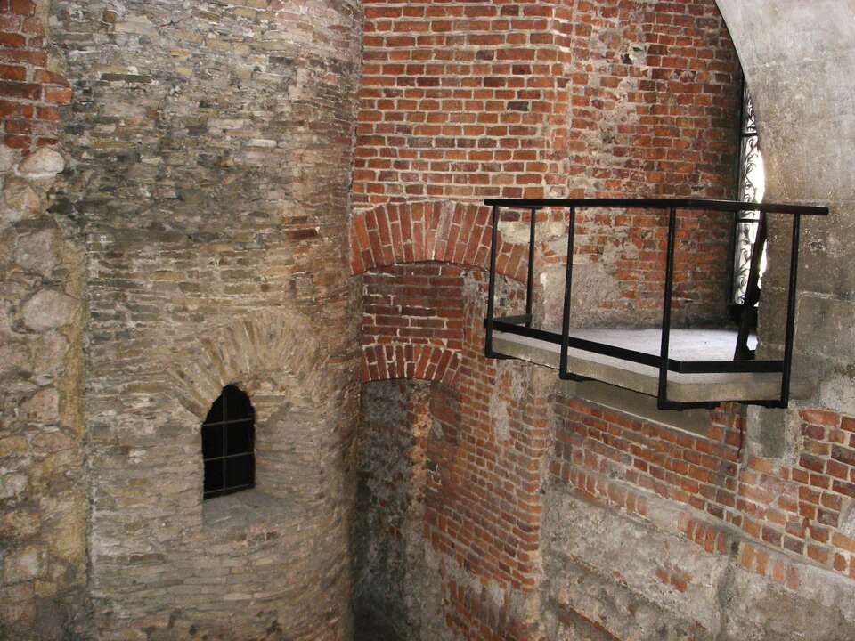Ilustracja przedstawia widok na fundamenty rotundy Najświętszej Marii Panny na Wawelu. Ukazuje fragment o surowym, kamienno‑ceglanym charakterze. Widoczne są grube, półkoliste ściany wykonane z nieregularnych kamieni oraz czerwonej cegły. W jednej ze ścian znajduje się niewielki, wąski otwór okienny o zaokrąglonym zwieńczeniu, zabezpieczony kratą. Po prawej stronie widoczna jest ceglana arkada oraz fragment podestu z metalową barierką, przeznaczonego dla zwiedzających. Różnice w materiałach i fakturach ścian wskazują na kolejne fazy budowy.