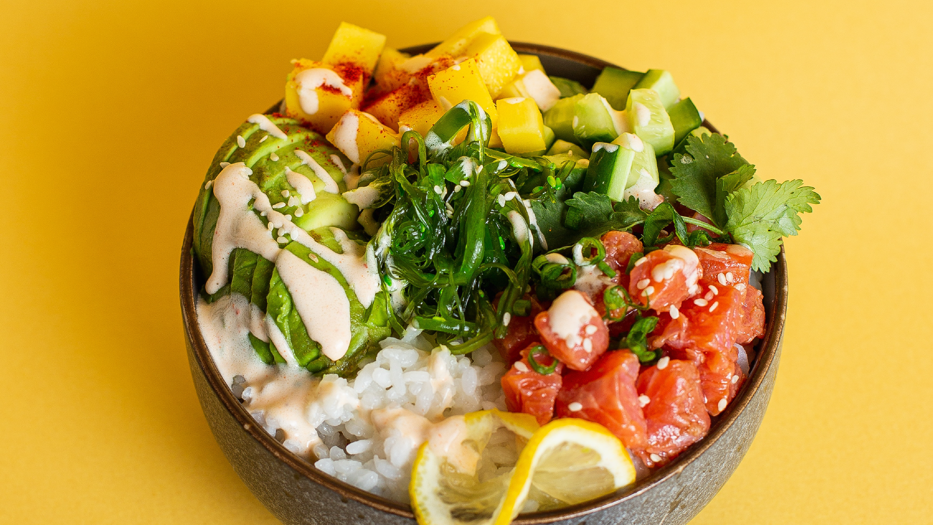 Zdjęcie przedstawia miseczkę, w której znajduje się sałatka. W sałatce znajdują się pokrojone w kostkę: pomidory, ogórki, awokado, jakiś owoc. Widoczny jest listek natki oraz plasterek cytryny, które ozdabiają sałatkę. Wszystkie składniki sałatki ułożone są na ryżu.