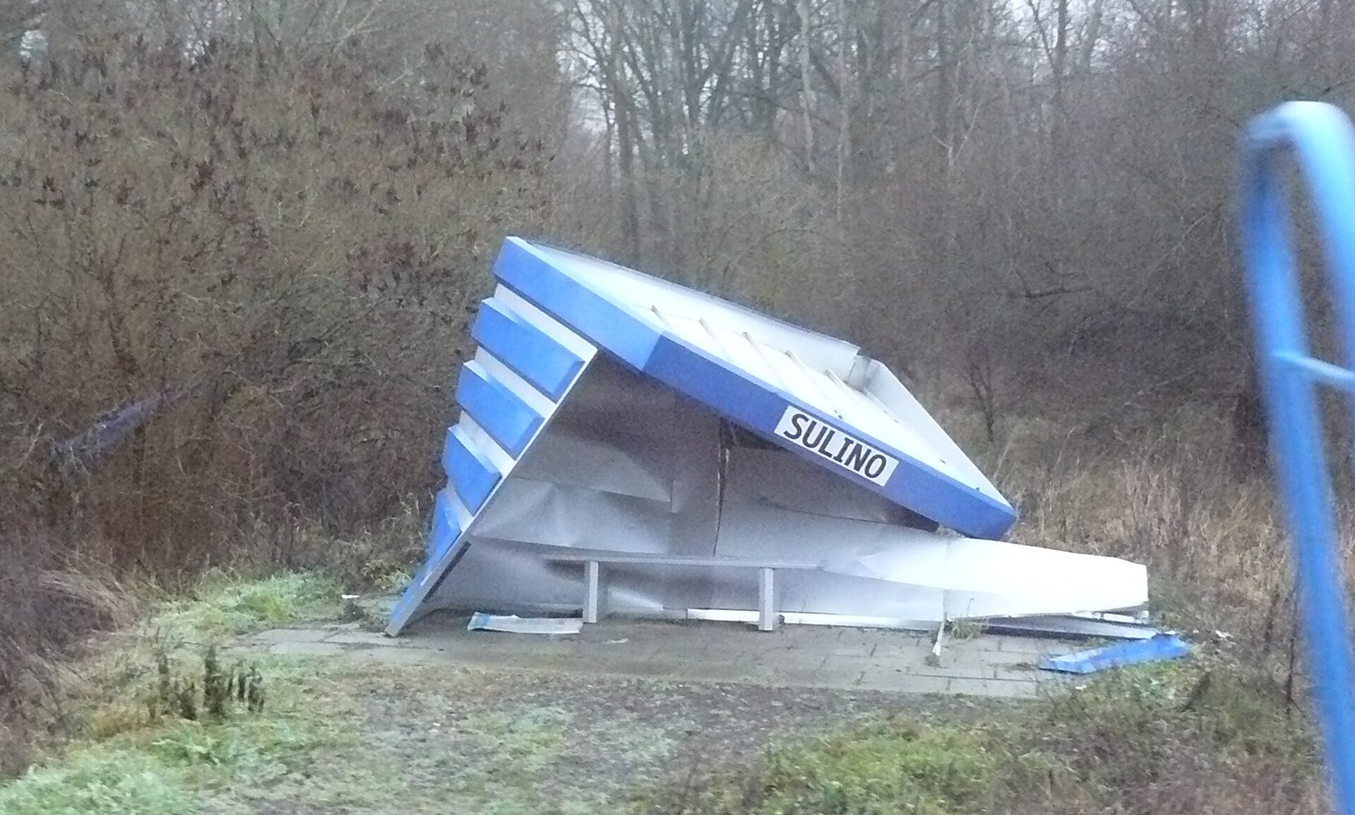 Zdjęcie przedstawia zniszczony przez wiatr przystanek. Na pierwszym planie znajduje się połamana wiata z ławką wewnątrz. W tle znajdują się liczne drzewa i krzewy.