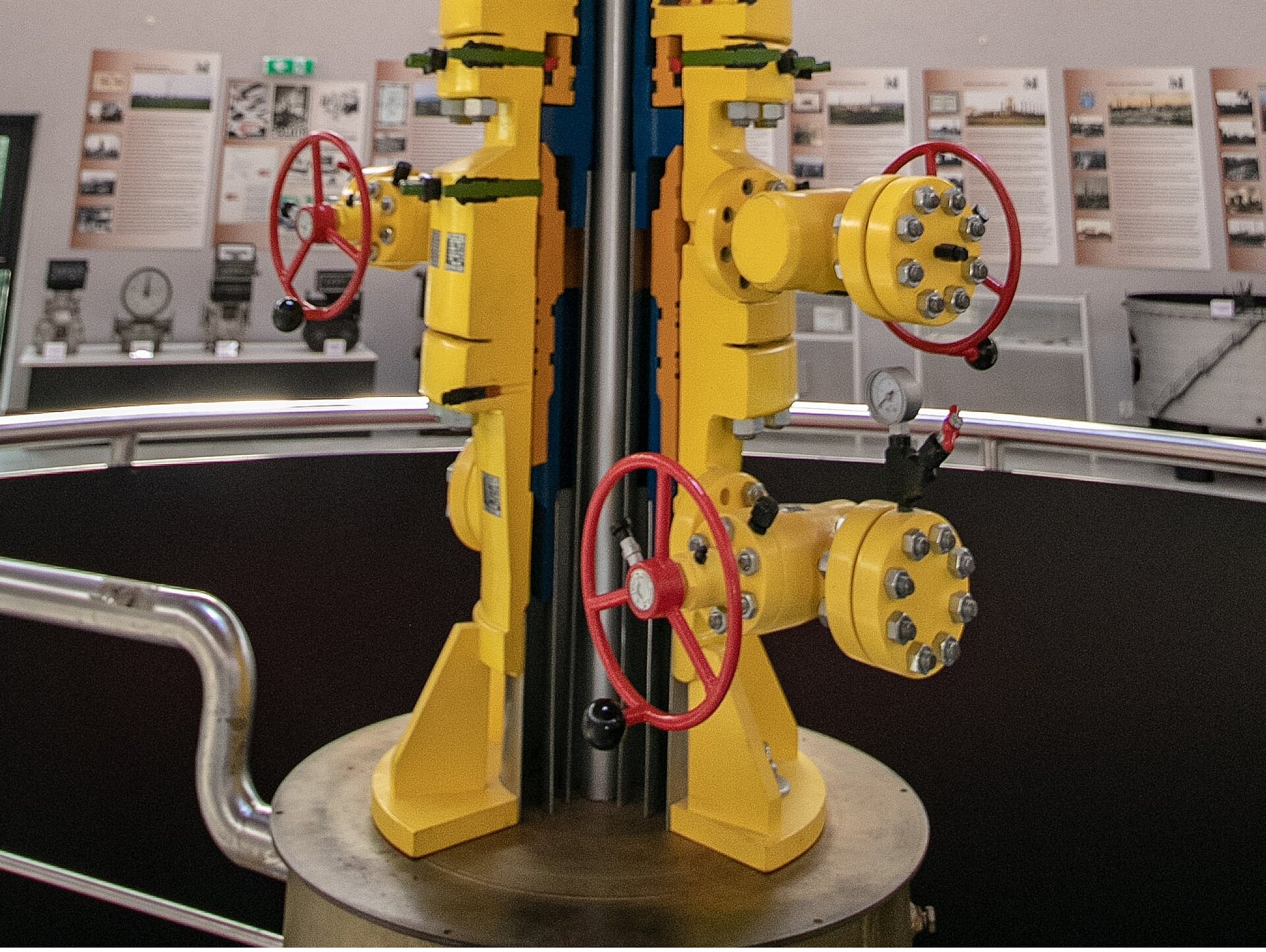 Na zdjęciu widać więźbę rurową kadłuby dolne i górne. Centralnym elementem grafiki jest głowica. Widoczna głowica jest pokazana w przekroju. Głowica składa się z cylindrycznych elementów połączonych ze sobą połączeniem kołnierzowym. W górnej części głowicy znajduje się element łącznikowy od którego odchodzą dwie odnogi. Obie odnogi zakończone są na końcu zaworami. Zawory są wyposażone w duże czerwone pokrętło. Przy podstawie głowica ma element łącznikowy od którego odchodzi jedna odnoga. Odnoga skierowana jest w lewo. Odnoga zakończona jest zaworem wyposażonym w duże czerwone pokrętło. W przekroju głowicy widać pionową stalową rurę. Rura znajduje się centrycznie.Widoczna na zdjęciu głowica umieszczona jest na podeście. Do podstawy podestu przymocowano rurę.W tle zdjęcia widać plansze i różne eksponaty.