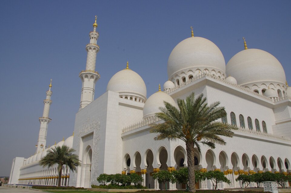 Ilustracja przedstawia meczet Szejka Zayeda bin Sultana Al. Nahyana w Abu Dhabi, Widoczny jest śnieżnobiały budynek, okalany arkadami kolumn z pozłacanymi elementami. Obiekt zwieńczony jest kopułami.