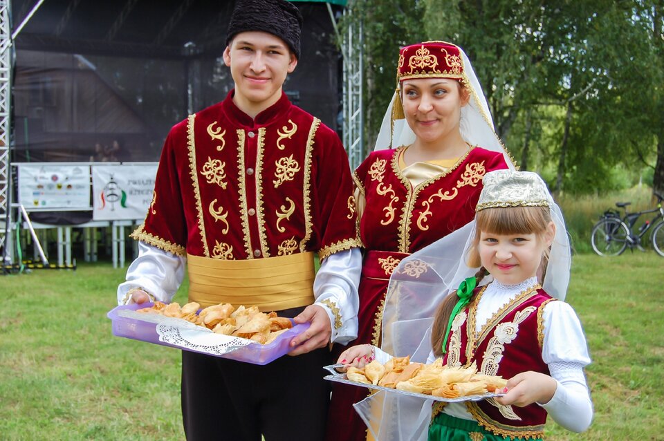 Ilustracja przedstawia grupę Tatarów - młodą kobietę, młodego mężczyznę i dziewczynkę, podczas V Festiwalu Kultury Tatarskiej. Wszyscy ubrani są w tradycyjne stroje ludowe. Mężczyzna i dziewczynka trzymają tacę ze specjałami kulinarnymi.