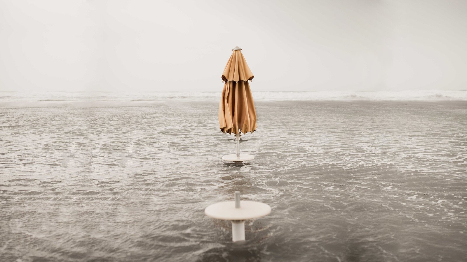 Zdjęcie przedstawia zalaną plażę. Z wody wystają jedynie dwa stoliki. Na jednym z nich widoczny jest złożony parasol.