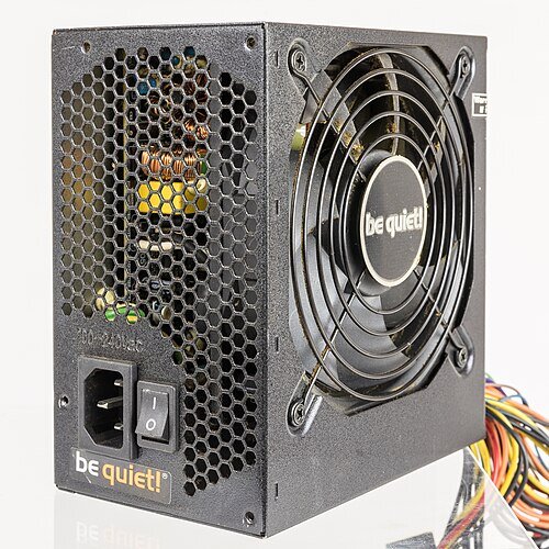 Obraz przedstawia zasilacz w kolorze czarnym, na jego bocznej ściance znajduje się duży wentylator chłodzący, zabezpieczony metalową kratką. Przedni panel wyposażony jest w złącze zasilania, włącznik oraz nazwę producenta. 