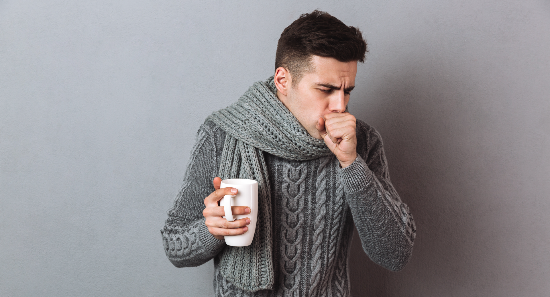 Ilustracja przedstawia kaszlącego mężczyznę w swetrze i szaliku, trzymającego w ręku duży kubek.