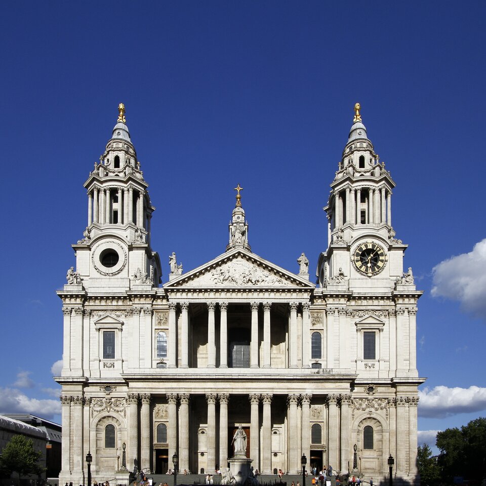 Klasycystyczna fasada katedry z dwiema symetrycznymi wieżami i centralnym portykiem z kolumnami. Na szczycie trójkątnego frontonu znajdują się rzeźby, a na jednej z wież widoczny jest zegar. Przed wejściem stoi pomnik na placu, a całość oświetla słońce na tle intensywnie niebieskiego nieba.