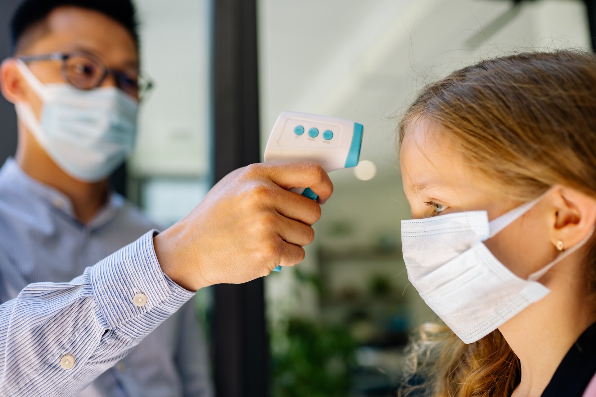 Zdjęcie przedstawia mężczyznę trzymającego w dłoni elektroniczny termometr bezdotykowy. Trzyma go przy czole stojącej obok dziewczynki. Obie osoby mają zasłonięte maseczkami chirurgicznymi nos i usta.