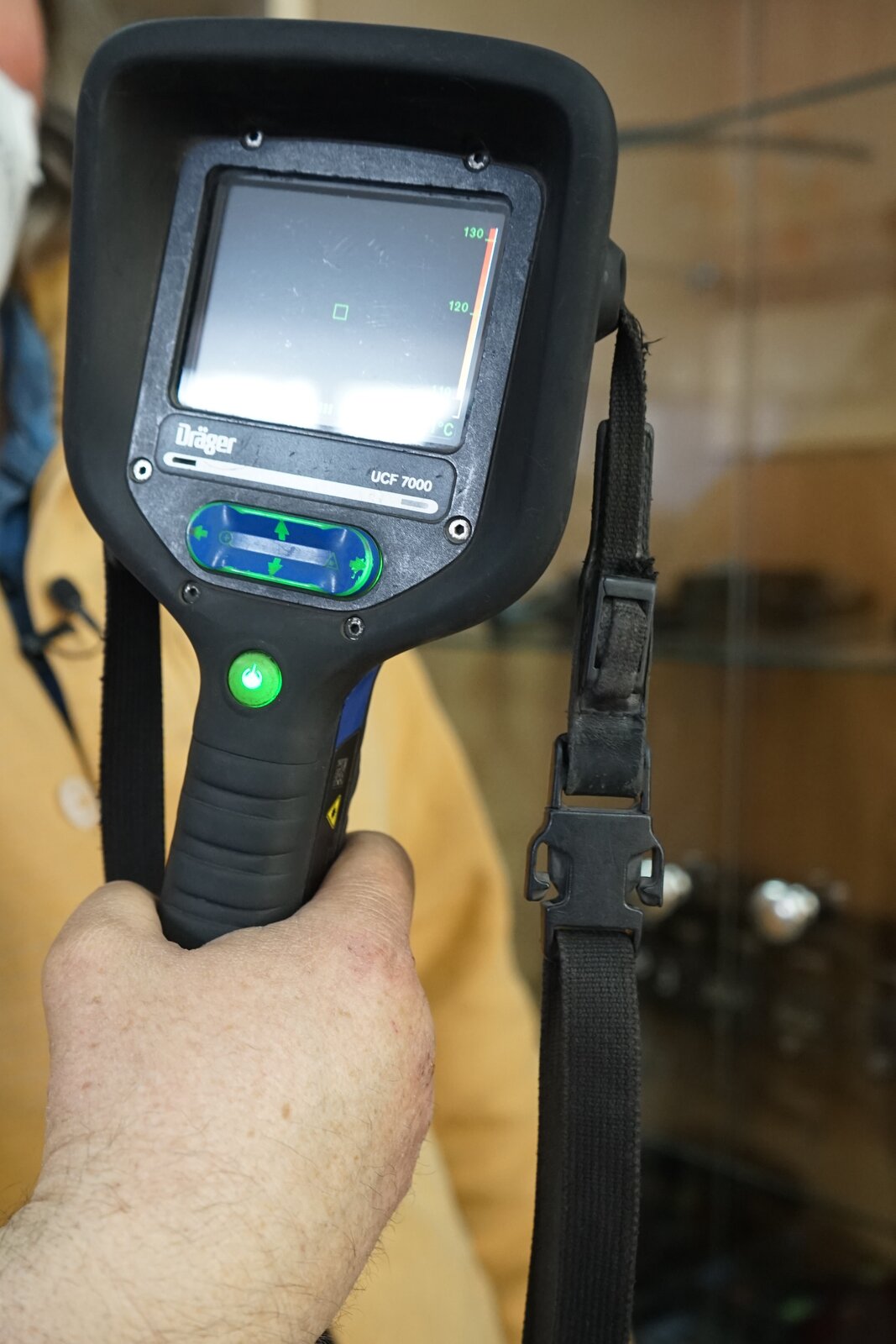 Zdjęcie przedstawia kamerę termowizyjną. Kamera osadzona jest na uchwycie pistoletowym. W górnej, prostopadłościennej części od strony osoby trzymającej urządzenie znajduje się prostokątny ekran. Pod nim zlokalizowane są przyciski. Do obudowy kamery przypięty jest pasek. Kamera jest prezentowana w ręce przez górnika. Górnik ubrany jest w strój ochronny.