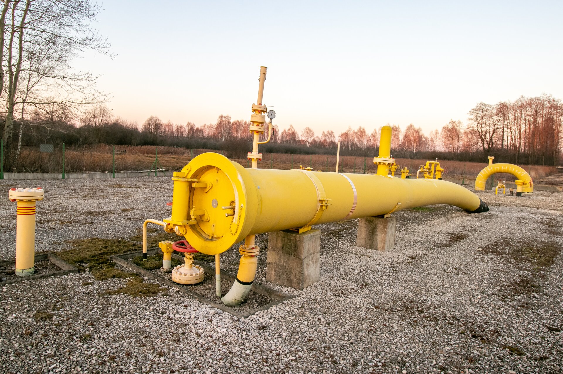 Na zdjęciu widać fragment rurociągu gazowego. Widoczny na zdjęciu fragment gazociągu jest stalową rurą w kolorze żółtym. Rura jest o przekroju okręgu. Na górze rurociągu widać dwa elementy rurowe które są prostopadłe do niego. Rurociąg posadowiony jest na betonowych prostopadłościennych klockach. Rurociąg zapada w prawą stronę wchodząc stopniowo pod ziemię. Początek rurociągu widoczny po lewej stronie zdjęcia jest zamknięty. Za rurociągiem w niewielkiej odległości widać układ rurociągów z zasuwami odcinającymi na gazociągu przesyłowym. Podłoże jest wyłożone żwirem. Cały układ jest widziany od przodu od strony rurociągu. Całość systemu znajduje się za ogrodzeniem. 